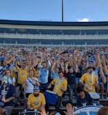A large group of enthusiastic sports fans, many wearing team jerseys in yellow and blue, are gathered in a stadium. They have their hands raised, showing excitement and support. Some fans hold scarves, while one person near the front is wearing a luchador mask. A few drums are visible, contributing to the lively atmosphere.