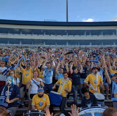 A large group of enthusiastic sports fans, many wearing team jerseys in yellow and blue, are gathered in a stadium. They have their hands raised, showing excitement and support. Some fans hold scarves, while one person near the front is wearing a luchador mask. A few drums are visible, contributing to the lively atmosphere.