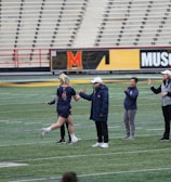 Several people are on a sports field with an empty bleacher in the background. One person is high-fiving a coach, while others stand nearby. They are wearing athletic clothing, hinting at a team sport environment. A sign with the text 'Muscle Milk' is visible in the background.