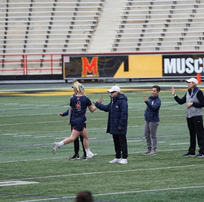 Several people are on a sports field with an empty bleacher in the background. One person is high-fiving a coach, while others stand nearby. They are wearing athletic clothing, hinting at a team sport environment. A sign with the text 'Muscle Milk' is visible in the background.