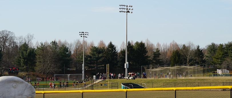 A sports field with a group of people engaging in athletic activities in the background. There is sports equipment like a soccer goal and a scoreboard visible. A chain-link fence runs across the foreground, and a statue or sculpture resembling an eagle is also seen. Trees and tall floodlights are positioned behind the field.