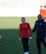 A group of people are gathered on a sports field. Some individuals are dressed in athletic gear, including a woman in a red sports jersey and a man beside her in a blue jacket, appearing to be in conversation. Additional people in similar athletic clothing are visible in the background.