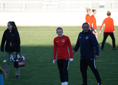 A group of people are gathered on a sports field. Some individuals are dressed in athletic gear, including a woman in a red sports jersey and a man beside her in a blue jacket, appearing to be in conversation. Additional people in similar athletic clothing are visible in the background.
