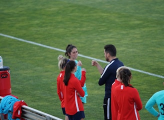 A group of people wearing sports attire are gathered on a grassy field. Some individuals are dressed in red jackets, while others are in turquoise and navy blue. A coach appears to be giving instructions. Sports equipment, such as water bottles and a bench with towels, can be seen nearby.