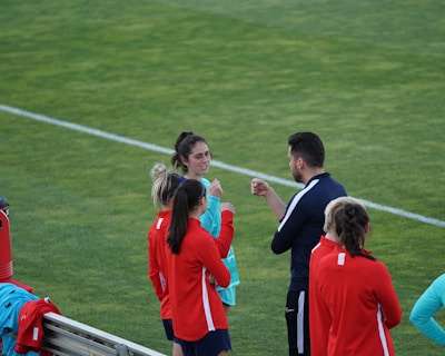 A group of people wearing sports attire are gathered on a grassy field. Some individuals are dressed in red jackets, while others are in turquoise and navy blue. A coach appears to be giving instructions. Sports equipment, such as water bottles and a bench with towels, can be seen nearby.