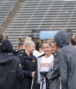 A group of people, likely athletes, are gathered together on a sports field. They are wearing black jackets with some individuals dressed in white sports uniforms underneath. A few people have their hair tied up in buns, and most are engaged in conversation. The background shows tiered stadium seats, suggesting the setting is an outdoor sports venue.
