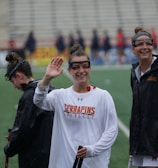 Several athletes in sports attire are on an outdoor sports field, interacting and smiling. One individual in the foreground waves with a friendly expression, while another stands beside her, also smiling. They wear protective goggles and gear associated with lacrosse. The background features indistinct figures in dark clothing against a backdrop of stadium seating.