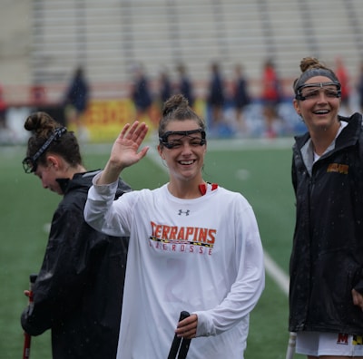 Several athletes in sports attire are on an outdoor sports field, interacting and smiling. One individual in the foreground waves with a friendly expression, while another stands beside her, also smiling. They wear protective goggles and gear associated with lacrosse. The background features indistinct figures in dark clothing against a backdrop of stadium seating.