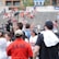 A lively gathering in an outdoor sports venue with a group of people, some wearing matching shirts labeled 'National Champions.' Several individuals are holding cameras and microphones, indicating a media presence. The background shows blurred spectators sitting on bleachers.