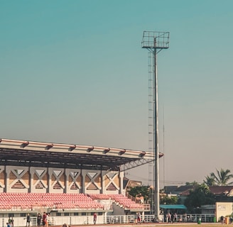 A large outdoor sports stadium with a green field and red spectator stands. A tall floodlight tower stands prominently beside the stands. A few people are walking and congregating near the seating area. The surrounding area includes some palm trees and a few buildings. The sky is clear and blue, suggesting a sunny day.