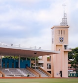 A sports field with a large seating area that includes blue and white bleachers. A prominent watchtower with a cross on top stands next to the seating area, displaying a crest. The area is surrounded by trees and has a cloudy sky overhead.