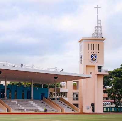 A sports field with a large seating area that includes blue and white bleachers. A prominent watchtower with a cross on top stands next to the seating area, displaying a crest. The area is surrounded by trees and has a cloudy sky overhead.