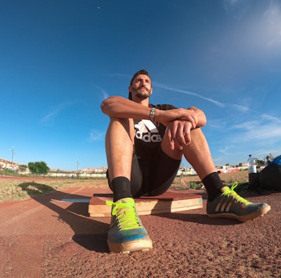 A person wearing a black athletic outfit is seated on a wooden platform in an outdoor sports field. The individual is relaxed, with arms resting on knees. The field is expansive with a clear blue sky and scattered clouds above. A brightly colored running shoe and athletic gear are visible, along with a water bottle and a blue backpack placed beside the person. Lampposts and residential buildings can be seen in the background.
