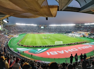 A large, brightly lit stadium filled with spectators. The field is green and well-maintained, with players actively engaged in a soccer match. The stands are crowded, and banners with writing are visible along the perimeter of the field. The atmosphere suggests excitement and energy.