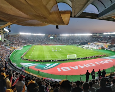A large, brightly lit stadium filled with spectators. The field is green and well-maintained, with players actively engaged in a soccer match. The stands are crowded, and banners with writing are visible along the perimeter of the field. The atmosphere suggests excitement and energy.