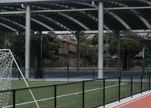 An outdoor sports facility featuring a running track and a soccer goal. The track is red, surrounded by a black metal fence, and the field is covered with artificial green turf. In the background, a basketball court is visible under a covered roof with metal supports. The sky is overcast, adding a moody atmosphere.