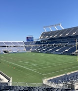 A large, empty sports stadium with blue seats surrounding a well-maintained green grass field. Bright sunlight creates clear shadows, and there are two goal posts on opposite ends of the field. A big electronic scoreboard displays some graphics, and the sky is clear and blue.
