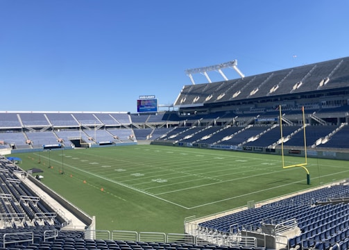 A large, empty sports stadium with blue seats surrounding a well-maintained green grass field. Bright sunlight creates clear shadows, and there are two goal posts on opposite ends of the field. A big electronic scoreboard displays some graphics, and the sky is clear and blue.