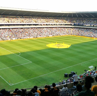 A large soccer stadium filled with a cheering crowd. The field is well-maintained with vibrant green grass, and players can be seen scattered on the pitch. The atmosphere is lively and bustling, typical of a sports event.